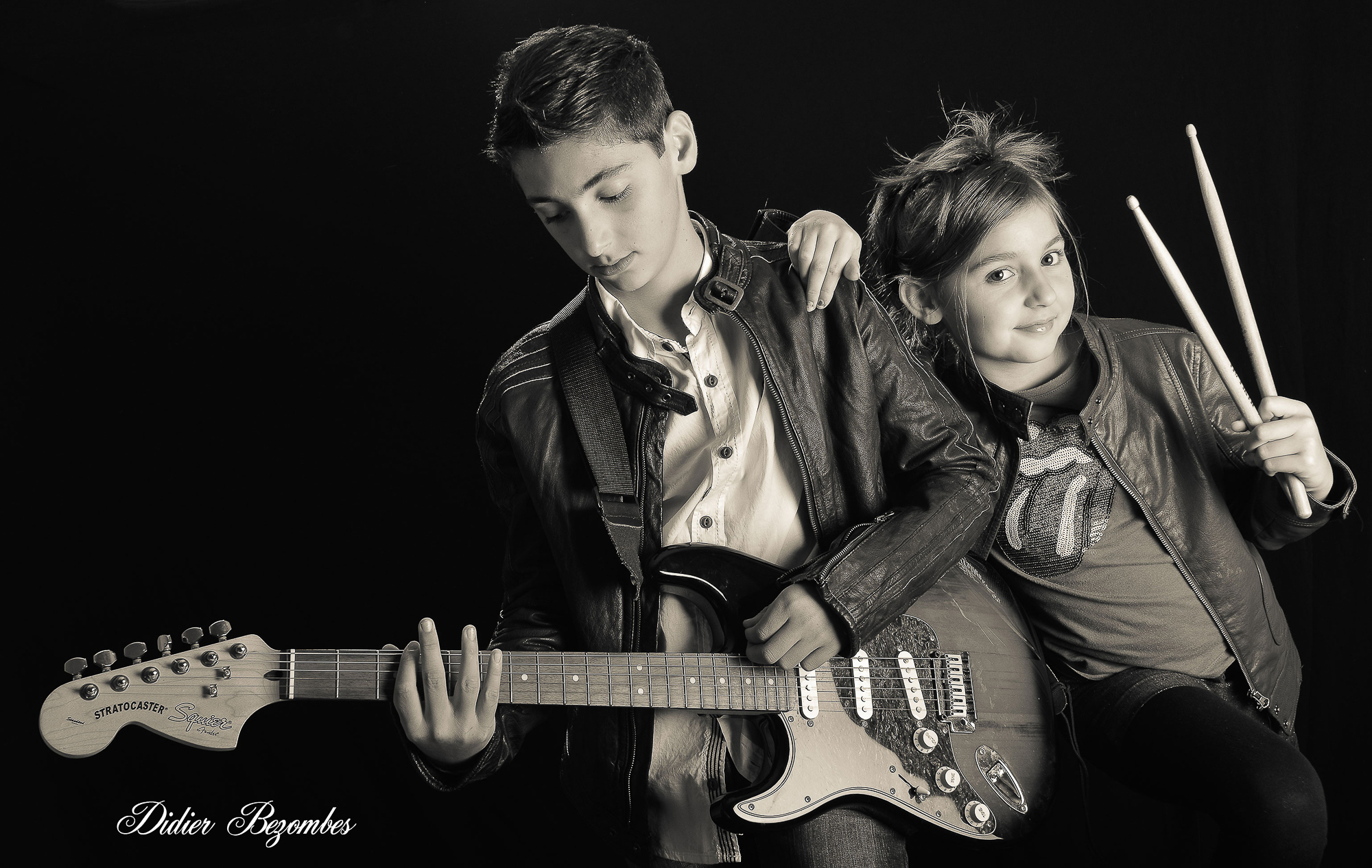 portrait-en-noir-et-blanc-photo-faite-en-studio-d'un-frère-avec-sa-soeur-le-garçon-a-une-guitar-electrique-et-la-fille-a-des-baguettes-de-tambour-photographe-de-portrait-Didier-Bezombes-Aveyron