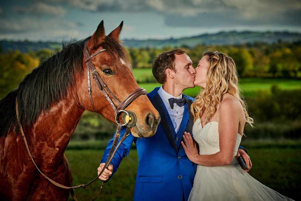 photo en couleur d'un jeune couple de marié qui s'embrasse c'est portrait d"eux leurs cheval alezan les regadent lui aussi est en portrait il a les oreilles droite le temps est ensoleillé c'est l'automne