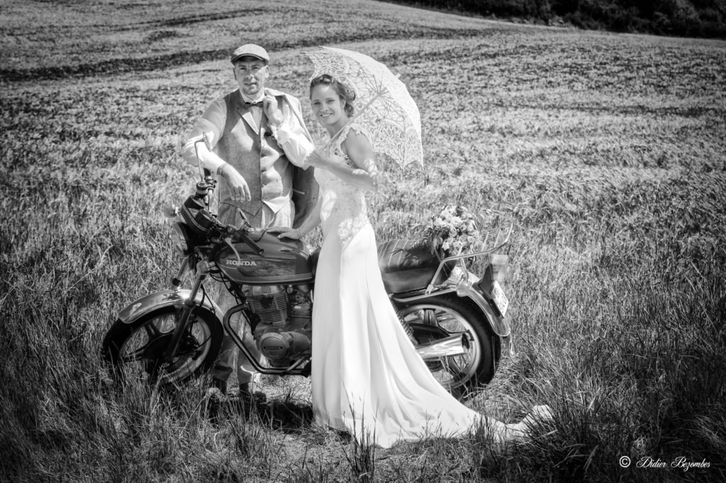 photos de mariage en noir et blanc il sont dans un champ avec une moto de marque Honda la mariée a une robe blanche en dentelles elle tiens une ombrelle le marié a un costume gris avec une casquette retro en aveyron sur le theme annees 1970 